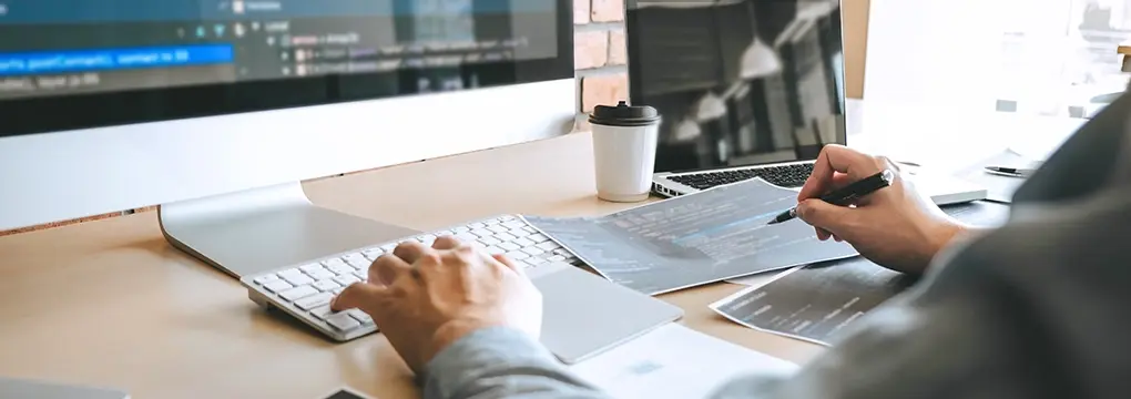 Writing with a pen and typing on a keyboard in front of a monitor.