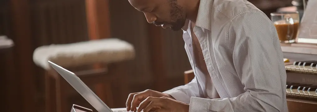 Man using a laptop in front of a piano with coffee.