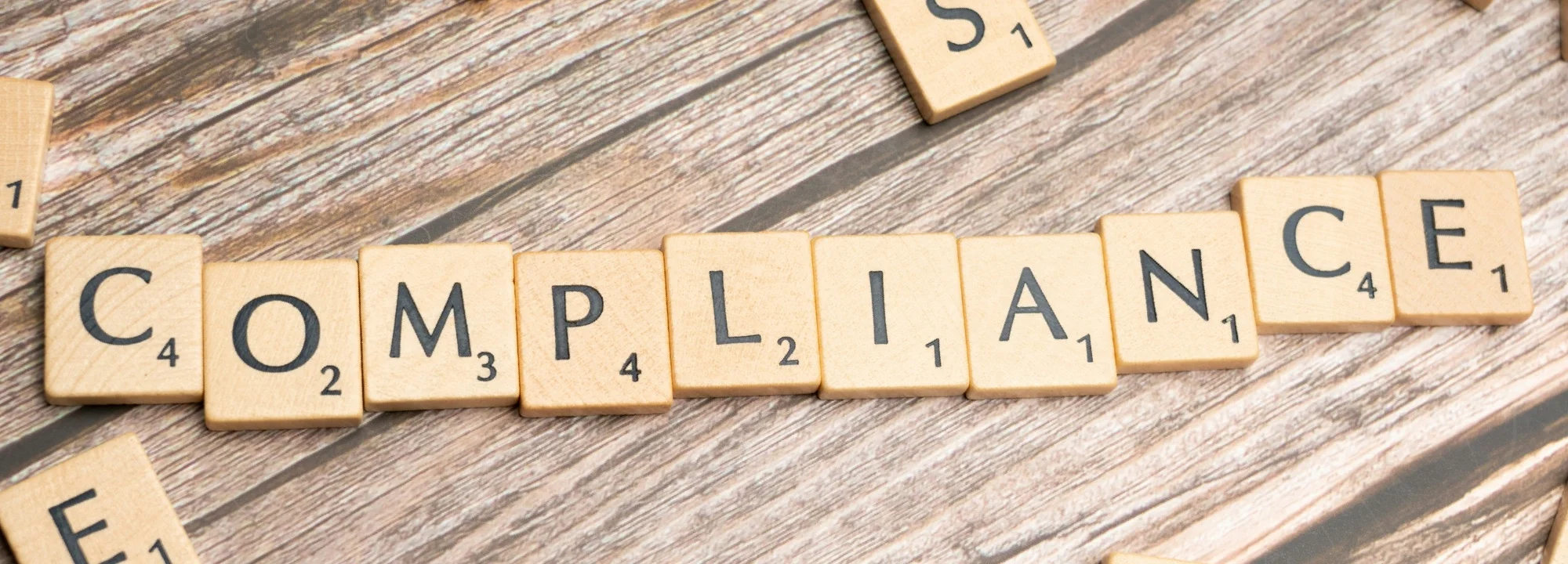 Scrabble letter tiles spelling COMPLIANCE on weathered gray wooden surface with scattered tiles nearby